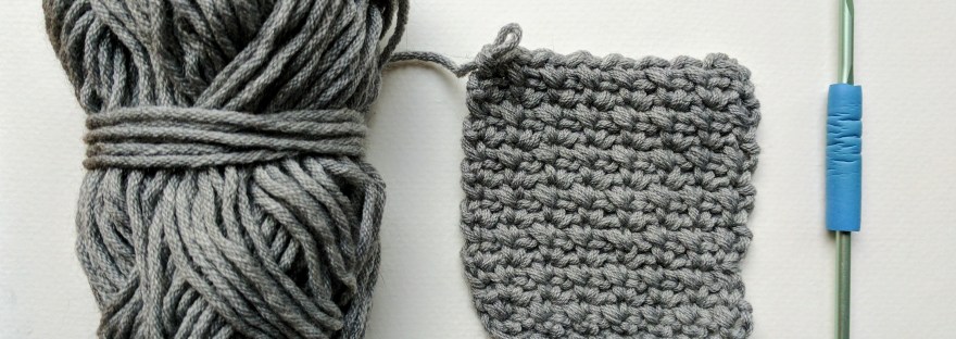 gray yarn skein, crocheted square in a textured stitch, and pale green crochet hook with blue grip, all on white background