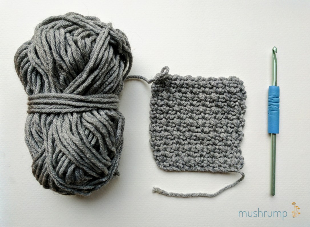 gray yarn skein, crocheted square in a textured stitch, and pale green crochet hook with blue grip, all on white background