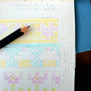 A portion of a graph-paper notebook with multi-colored designs in it: hearts, cupcakes, and flowers. A black pencil is resting on the notebook, and some pattern words are at the top.
