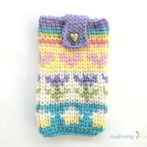 A crocheted pouch on a white background with a logo in the bottom right corner that says Mushrump and has yellow mushrooms next to it. The pouch looks knit and has pastel hearts, flowers, cupcakes, and a rainbow on it. There is a metal heart button near the top.